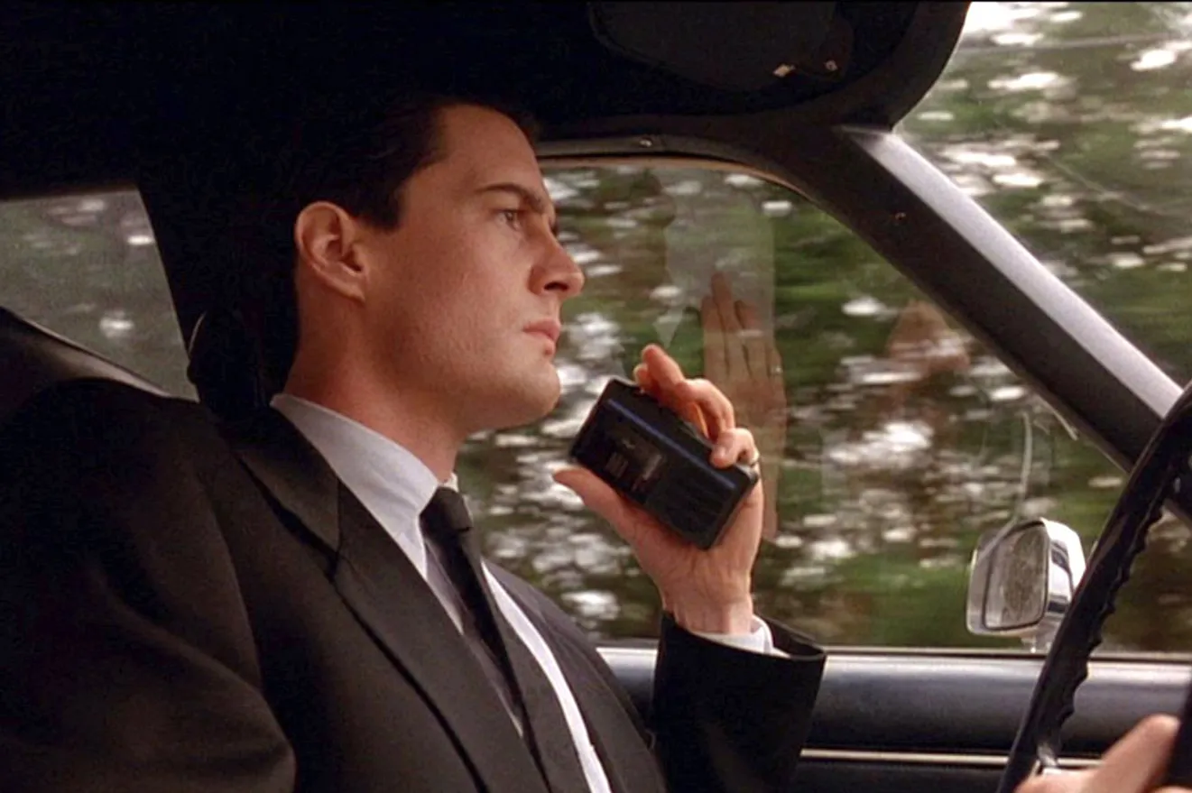 Agent dale cooper is driving a car while recording a note on a hand-held recorder.